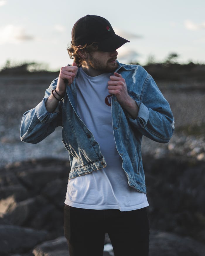 why-choose-us Man in a denim jacket and cap on a rocky outdoors setting, showcasing casual fashion.