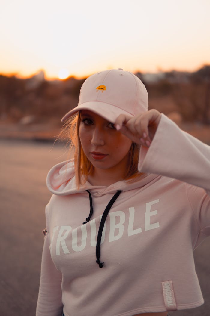 gallery-3 Young woman wearing a pink cap and hoodie, posing outdoors at sunset.