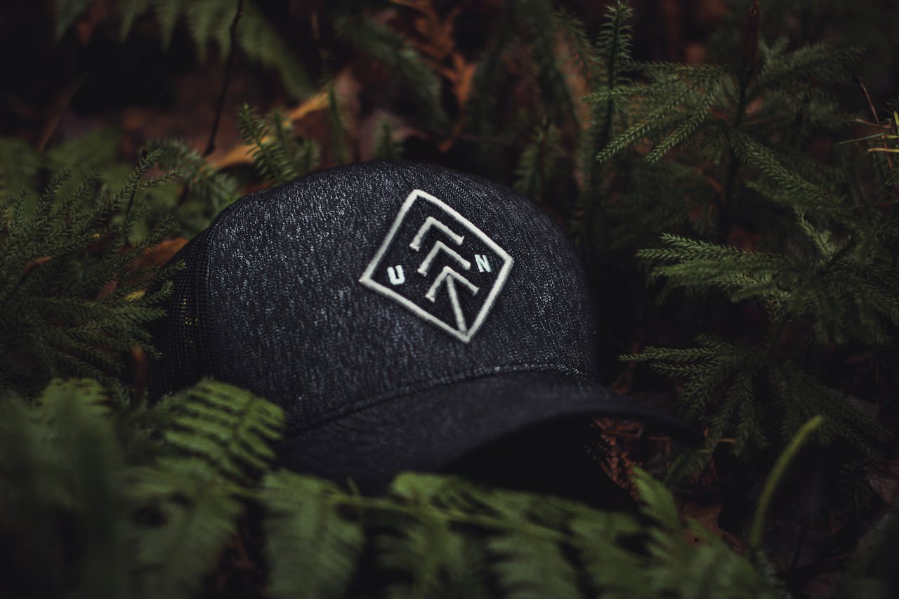 hero-img-01 Close-up of a dark baseball cap with an embroidered design nestled among green ferns, creating a nature-themed concept.